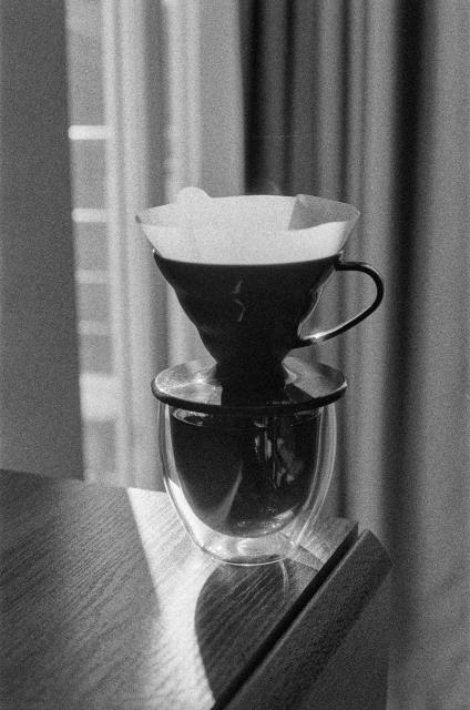 The image shows a black-and-white scene featuring a pour-over coffee setup. It includes a cone-shaped coffee dripper placed on top of a transparent double-walled glass. A paper filter is inserted into the dripper, which sits on a wooden surface with soft natural light illuminating part of the scene. The light creates shadows on the table, and the overall atmosphere is calm and minimalist. The grainy texture gives the photograph a vintage or film-like quality, enhancing its timeless feel.