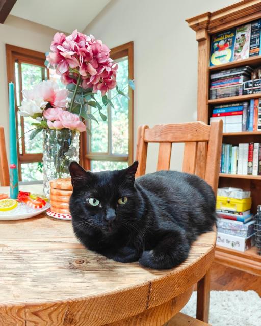 A black cat laying on a wooden table next to a vase of flowers.