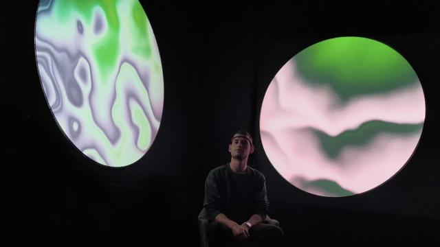 Photo of a man sitting down, wearing an EEG reader, in front of two circular visualizations. Black background.