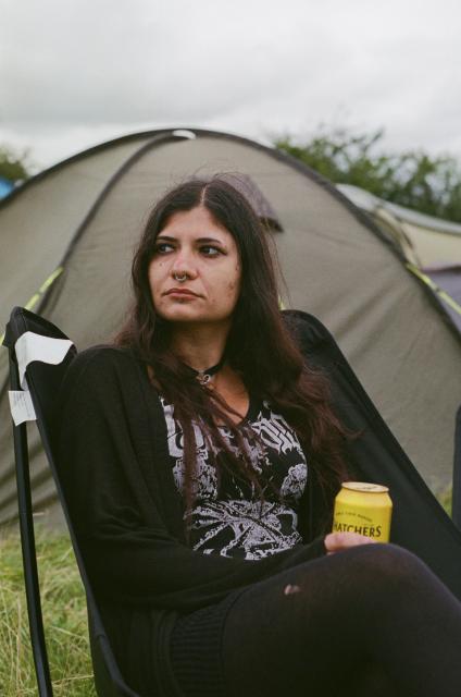 A woman with a nose ring and dark hair that's been dyed slightly purple, wearing black clothes and a choker, holding a can of cider while sitting in a camping chair on a field of grass. In the background, there are camping tents.