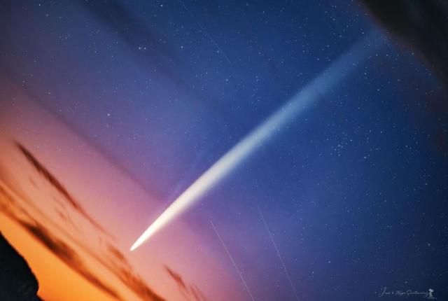 A starry sky is pictured with a long bright streak running diagonally from the lower left to the upper right. The lower left part of the sky sky orange sprinkled with a few dark clouds.