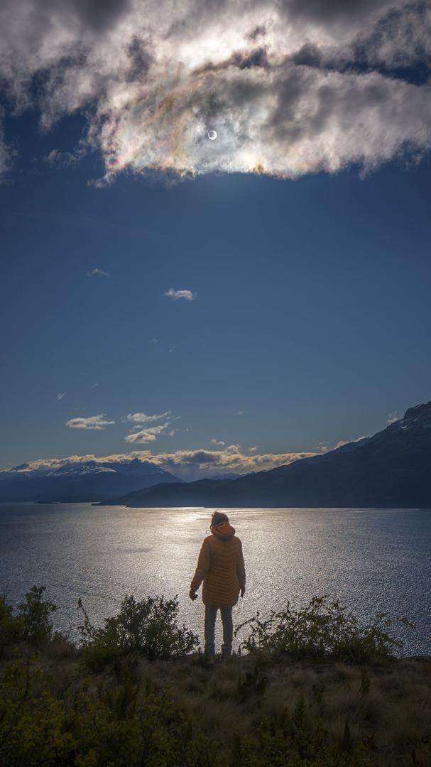 A person stands looking over a lake. High in a partly cloudy sky is the Sun. A close look at the Sun will show that there is a dark spot in the center -- the Moon during an annular eclipse.