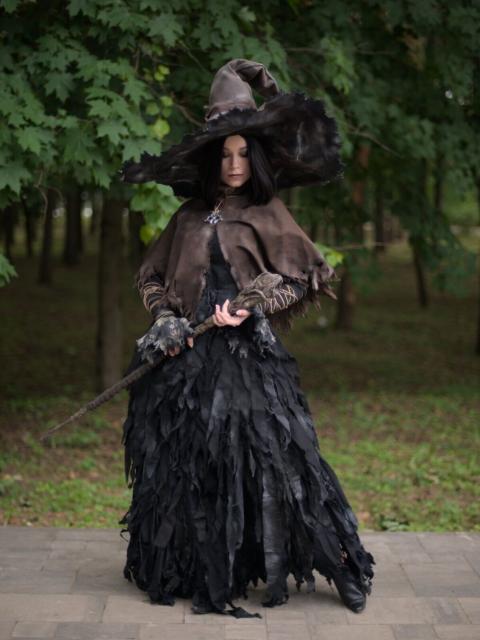 A portrait of a young woman wearing black pointed wide-brimmed hat, brown leather cape, long black dress with lower part made of different little pieces of cloth, and black leather boots. She has relatively short staff that she's holding in both hands. Her left leg is put a bit to her left. She looks down and to her left, her ayes are almost closed. She has shoulders-length black hair.