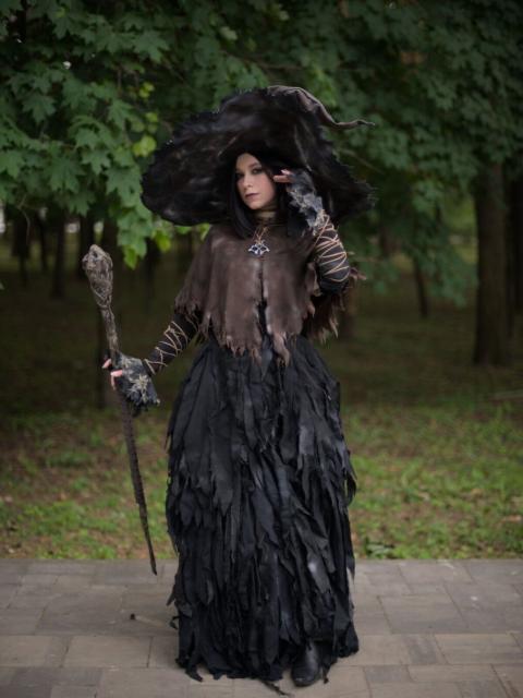 A portrait of a young woman wearing black pointed wide-brimmed hat, brown leather cape, long black dress with lower part made of different little pieces of cloth, and black leather boots. She has shoulders-length black hair. She has relatively short staff that she's holding in right hand. Her left leg is put a bit forward. Her left hand is near her face, as if she's adjusting her hair. Her head is tilted slightly to her right and back. She looks straight to the camera.