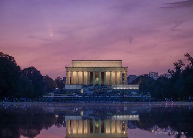 The Lincoln Memorial monument in Washington, DC, USA is pictured from afar. Behind the monument is a sunset-colored pink sky. In the sky, on the upper left, is a white streak that is a comet.