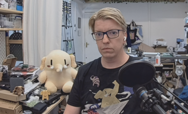 A picture of the author, a white male in his late 40s with blond hair and glasses, looking into the camera. He is sitting in a maker studio with white walls and a variety of things surrounding him. He is wearing a black t-shirt with a Mastodon illustration in a cartoony style, and there is a large plush Mastodon toy over his right shoulder. There is a microphone and pop shield in the foreground.