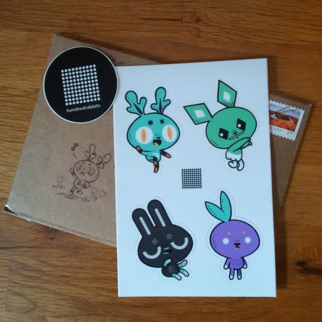 A photo of a set of stickers, featuring four whimsical rabbit characters -- including a purple turnip rabbit -- sitting on top of the small cardboard envelope they came in, which also features a singing rabbit drawn in black pen, running hopping through a field of dandelions. They're awesome, trust me.