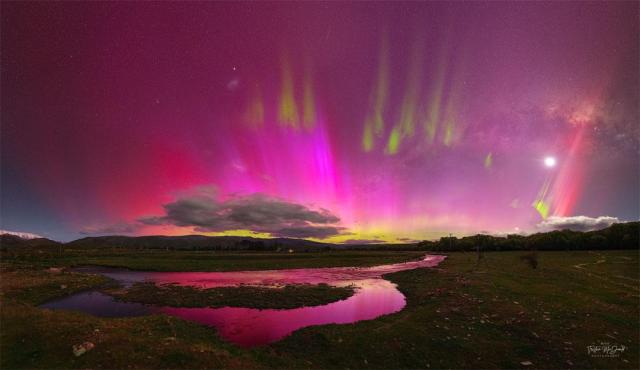 A night sky is shown that appears mostly red due to pervasive aurora. In the foreground is covered by watery grasslands. Clouds are visible above the horizon. Thin green aurora are visible toward the top of the frame. In the background one can find the Moon, the LMC, SMC, Venus, a meteor, and the band of our Milky Way galaxy.