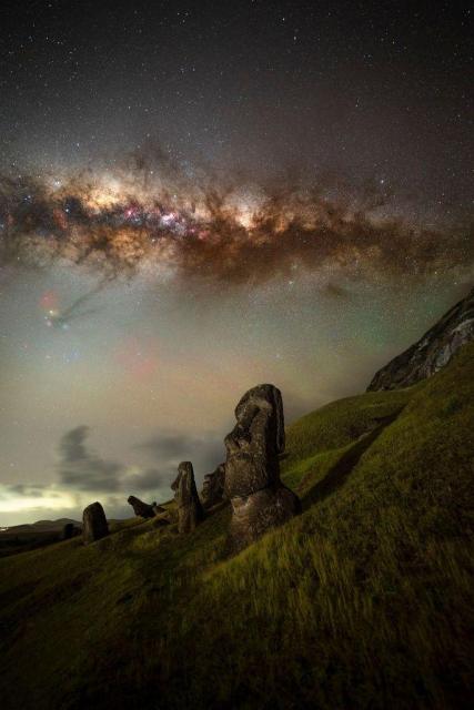 The Milky Way is a thick, dusty band almost parallel to the bottom of this image. In the foreground are at least four Moai statues on the hill side of Easter Island. 