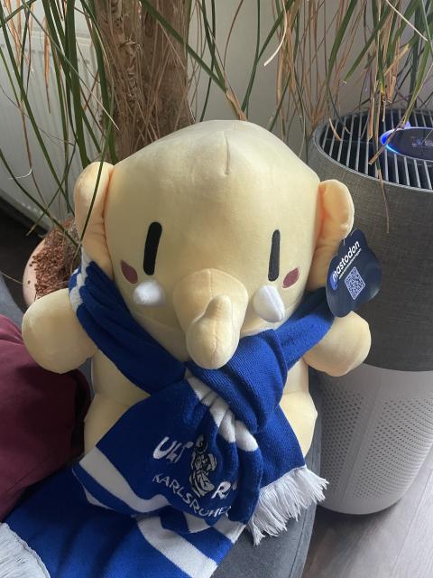 A plushtodon wearing a blue and white ksc ultras scarf sits in front of a decorative plant and a small speaker. The scarf features the logo of a sports team, and the elephant has a cheerful expression.