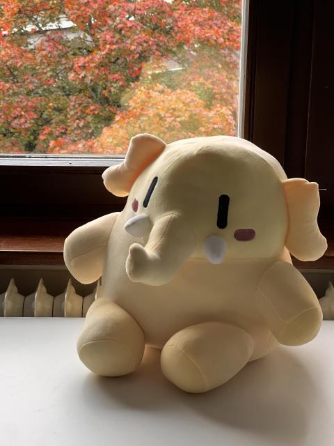 mastodon plushie sitting on a white table infront of a window. there are the leaves of a tree outside