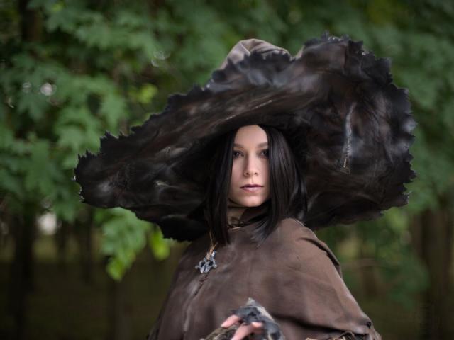 A chest-height portrait of a young woman wearing black pointed wide-brimmed hat and brown leather cape. She wears a pendant made of gray metal, resembling very stylized skull. She has shoulders-length black hair. Her head is turned slightly to her left. She looks attentively at something behind the camera in the right side of the frame.