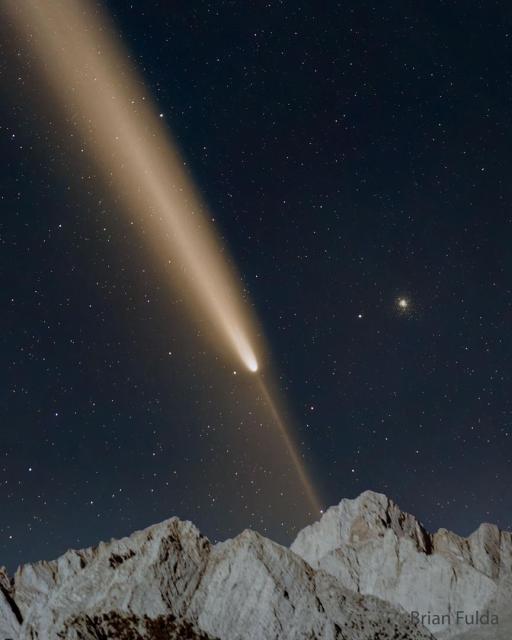 A starfield is shown with a bright comet. The main tail of the comet points diagonally to the upper left, while a thin anti-tail points to the lower right. Mountain peaks are visible at the bottom in the foreground.