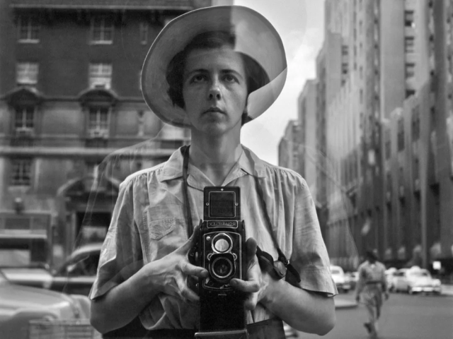 Self portrait of Vivian Maier taken in New York in 1954. In a black-and-white image, she is holding a camera up to a window. There is a city street behind her and cars of the era, with one person, blurred, walking on her left-hand side. 