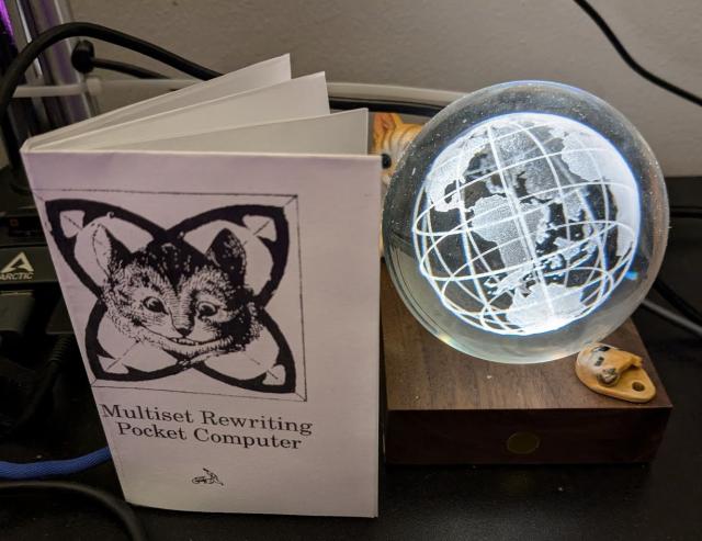 a picture of a zine entitled "Multiset Rewriting Pocket Computer". the zine is standing besides a glass, illuminated globe. there's a smiling cat on the cover of the zine, and behind the globe, there's a statuette of a cat, facing the zine.