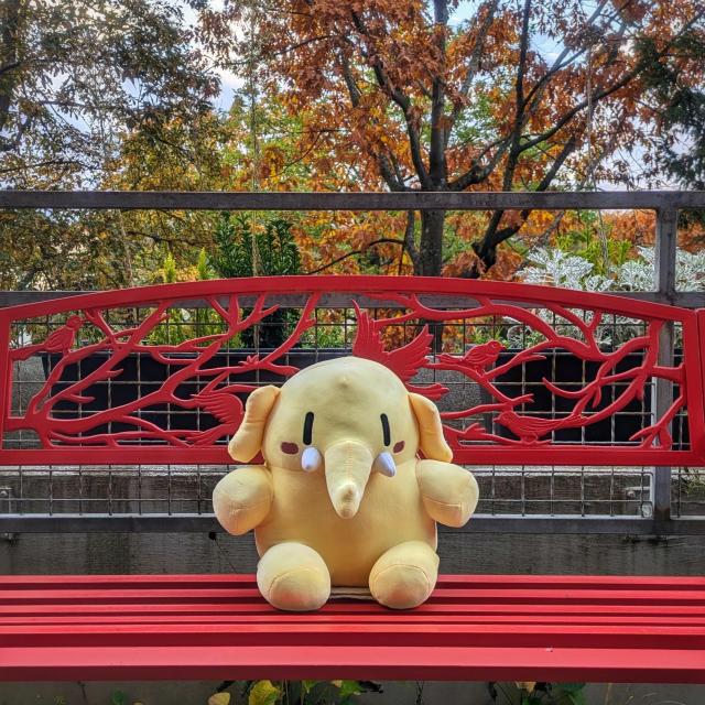 The Mastodon plushie is sitting on a red bench with colorful autumn trees in the background.