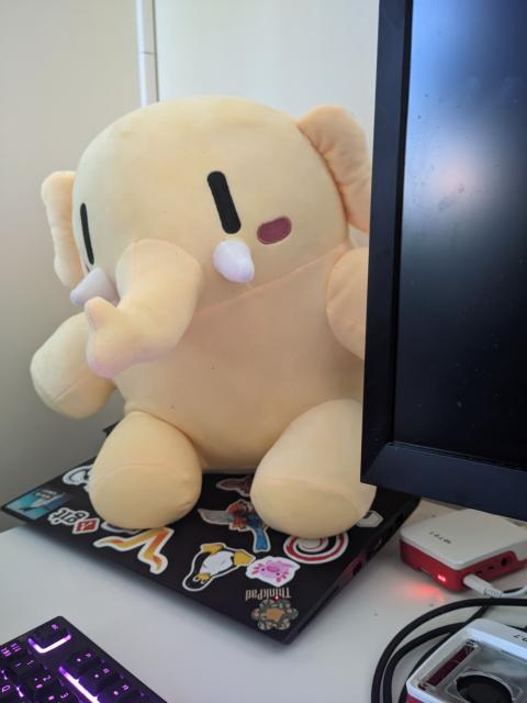 A yellow Mastodon plushie sitting on my laptop next to a couple of Raspberry Pis, a keyboard, and a blank screen. It looks like they're leaning out from behind the screen.