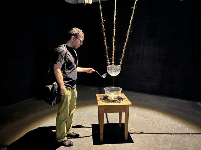 a handblown glass bowl hangs from above via three jute ropes, brightly illuminated against a dark background. A person is holding a ladle and watching as water drips down into a second bowl on a small wooden table surrounded by dried seaweed.

