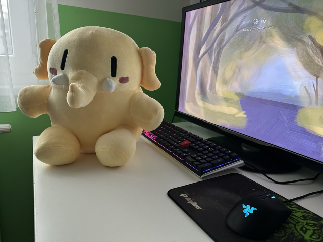 The mastodon plushie sitting in front of a PC, eagerly waiting to be cuddled.