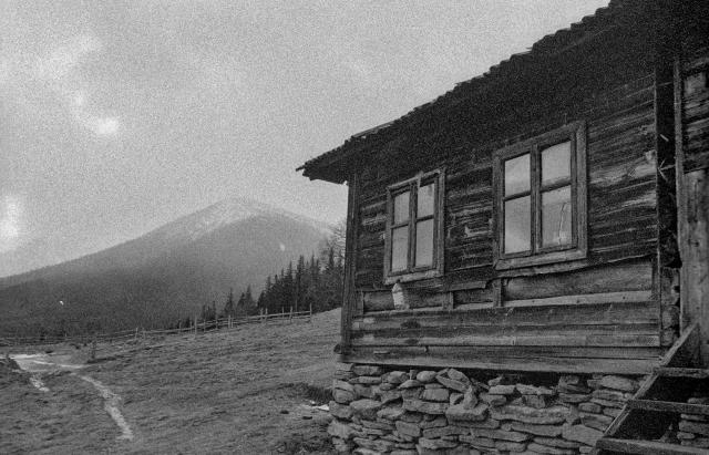 The image depicts a rustic, wooden cabin in a rural setting, shot in black and white. The weathered structure has two large windows with simple frames, and the walls are made of wooden planks, sitting atop a stone foundation. A small staircase leads up to the side of the cabin. In the background, a mountain looms, its peak partially shrouded by mist or clouds. The terrain around the cabin is hilly, with a winding path that leads into the distance, bordered by a fence. The scene has a rough, grainy texture, giving it a moody, atmospheric feel, suggesting it was shot on high-ISO film. 

