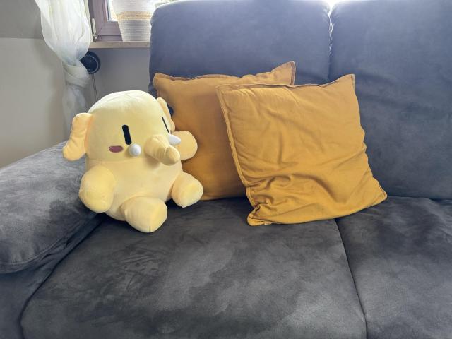 A cozy seating area featuring a gray sofa, two mustard yellow cushions, and a cute yellow mastodon plush toy. Natural light filters in from a nearby window.