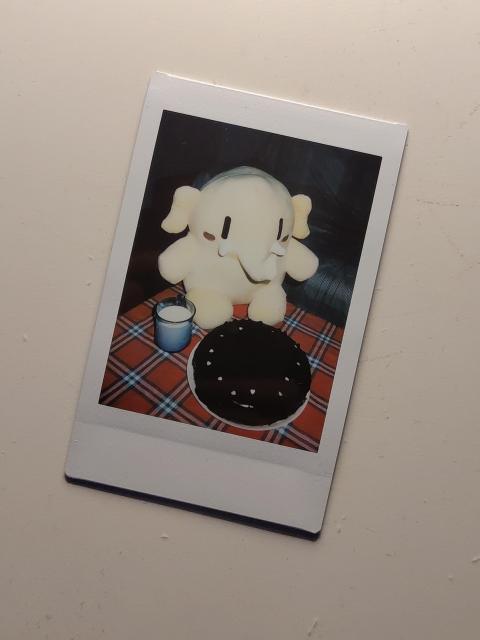 Instant photography of the Mastodon plush with a chocolate cake and a glass of milk.