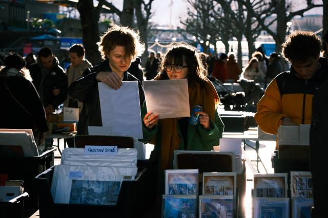 A cinematic colour photo of a young couple looking at prints of literary illustrations at the book market beneath Waterloo Bridge, on London's South Bank. The sun is behind them and is lighting up their hair from behind, and is also casting reflected light from the prints they're holding onto their faces. A number of prints can be seen propped up on the table in front of them, facing the camera, including a large one by Gustave Doré.