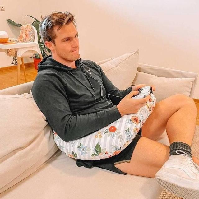 A guy using a nursing pillow while going from the couch. He is holding a Dualsense (Playstation) controller while his arms are supported by the nursing pillow.