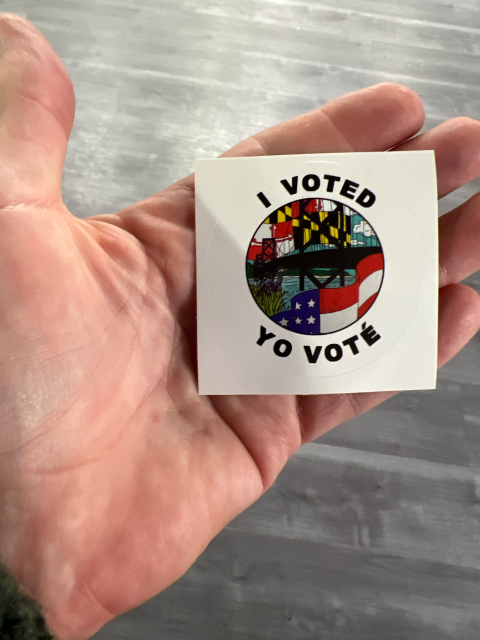 The hand of an older white man holds a round sticker with the United States and Maryland flags, and text that reads (in English and Spanish) "I voted"