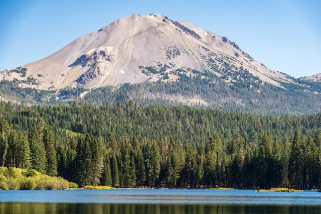 Behind a lake with a tiny little paddleboarder, a forest and mountain loom amidst blue sky. [Fuji X-T5 / Tamron 18-300]