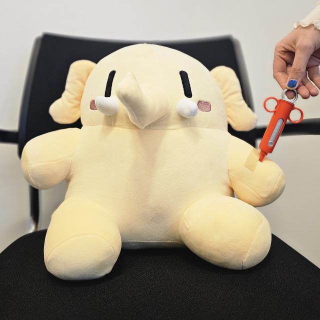 Ein Mastodon Plüschtier sitzt auf einem Bürostuhl in Richtung Kamera, von rechts hält ein menschlicher Arm eine Spielspritze so, als würde das Plüschtier eine Impfung in den Arm gespritzt bekommen.