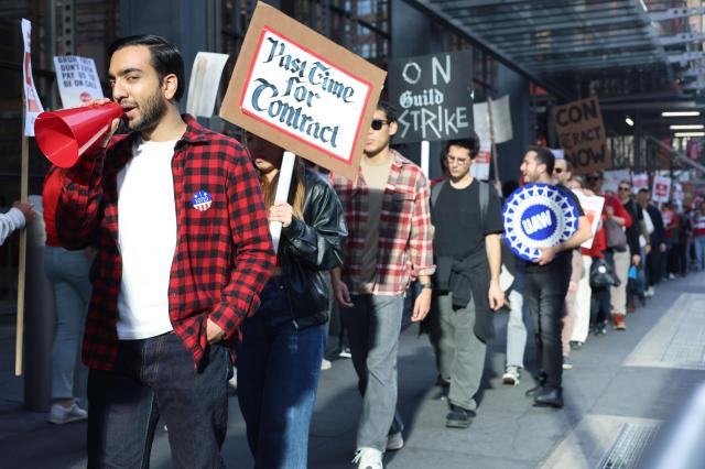 A photo of the picket line outside of the New York Times headquarters at 620 8th Ave in Midtown Manhattan. The line stretches beyond the far right edge of the frame. One worker at the far left wearing a checkered red and black longsleeve shirt is holding a bullhorn, and the person behind them is holding a “Past Time for Contract” sign written in blackletter script.