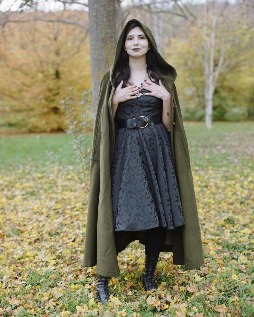 A beautiful woman with black hair, wearing black lipstick, a black dress and an olive wool coat as well as a necklace in the shape of a moth, standing in front of a green and yellow landscape with lots of yellow leaves on the grass.