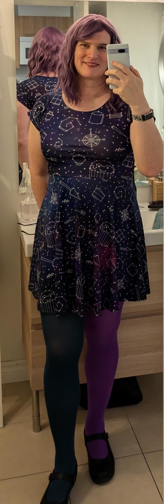 Mirror selfie. I have shoulder-length wavy purple hair and am wearing a purple dress with a white constellation pattern, featuring stars and various geometric shapes. I'm also wearing mismatched tights—one leg in dark teal and the other in purple—and black Mary Jane shoes. 