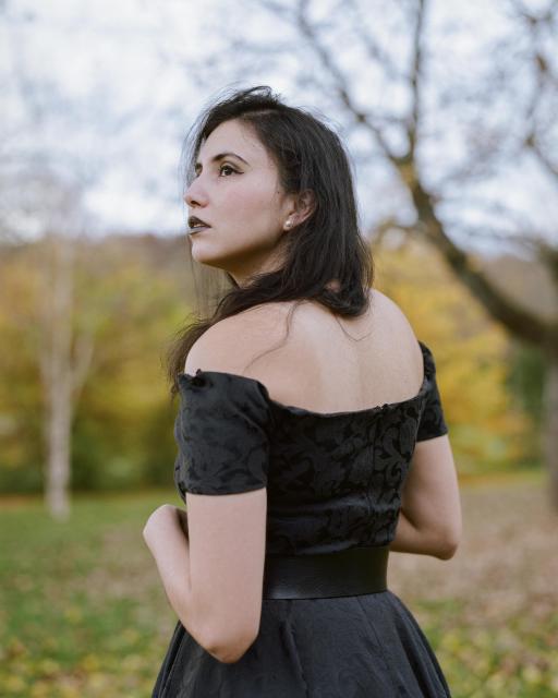 A beautiful woman with black hair, wearing pearl earrings, black lipstick, and a black off-shoulder dress, in front of a green and yellow autumnal landscape.