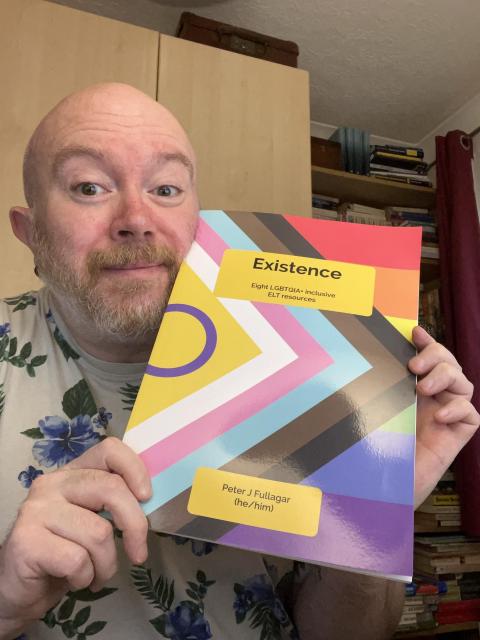 Peter, a cis white bald man with a beard holds up Existence, a book with the intersex inclusive pride progress flag as cover. He wears a floral T-shirt and is in a small office with a wardrobe and bookcase behind him. 