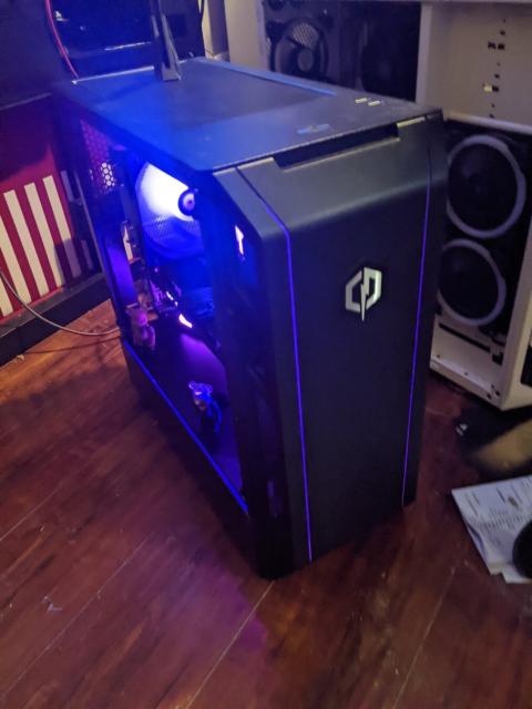 Photo of a desktop PC in a bedroom. PC is black with purple LED lights and sits on a wooden floor. Several fans are visible inside the case and are similarly illuminated