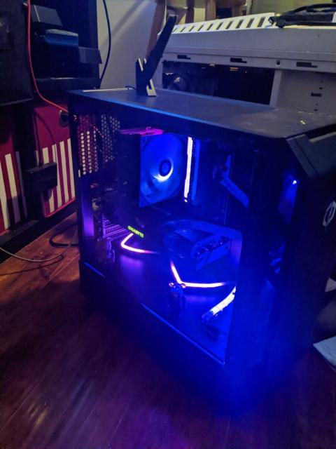 Photo of a desktop PC in a bedroom. PC is black with purple LED lights and sits on a wooden floor. Several fans are visible inside the case and are similarly illuminated