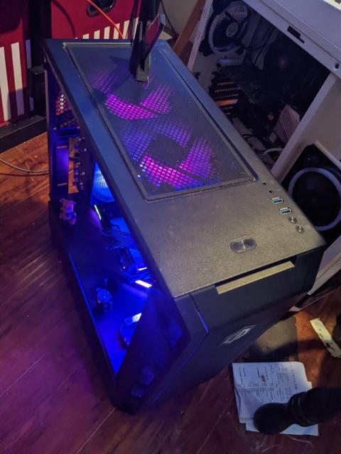 Photo of a desktop PC in a bedroom. PC is black with purple LED lights and sits on a wooden floor. Several fans are visible inside the case and are similarly illuminated