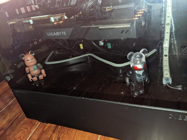 Photo of a desktop PC in a bedroom. PC is black with purple LED lights and sits on a wooden floor. an LED strip wraps around two figurines inside the case