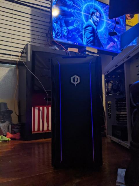 Photo of a desktop PC in a bedroom. PC is black with purple LED lights and sits on a wooden floor. Several fans are visible inside the case and are similarly illuminated

Behind the PC a stylized wallpaper of John Wicke, a white man in a suit with a gun, is displayed on a TV