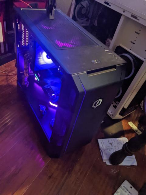 Photo of a desktop PC in a bedroom. PC is black with purple LED lights and sits on a wooden floor. Several fans are visible inside the case and are similarly illuminated