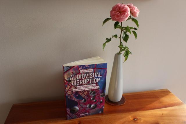 Book titled "Audiovisual Disruption: Post-Digital Aesthetics in Contemporary Audiovisual Arts" stands on a wooden surface next to a vase with two roses.