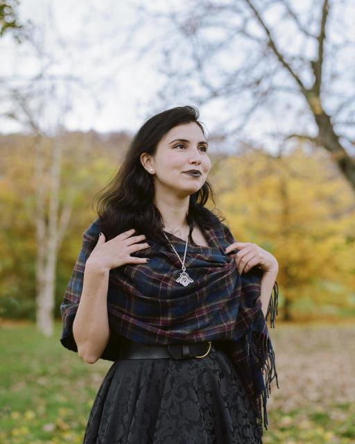 A beautiful woman with black hair, wearing pearl earrings, black lipstick, a silver moth necklace that is unfortunately hanging the wrong way around, and a black dress covered by a plaid shawl, in front of a green and yellow autumnal landscape.