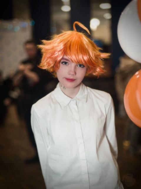 A waist-height portrait of a young woman wearing long white shirt with long sleeves. Her medium-length bright-orange hair is styled, but in some places sticking in different directions. On her neck there is a tattoo consisting of digits only, and just two last blurred digits are really visible. She holds her hands behind her back. She looks directly into the camera with very light smile.