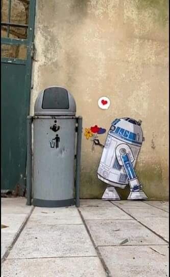 Street art mural depicting R2-D2, the iconic Star Wars droid, offering a bouquet of flowers to a trash can painted to resemble a companion. A small red heart hovers above R2-D2, adding a touch of charm and humor to the piece. The artwork, set against an urban wall, playfully reimagines the characters in a heartwarming scene that brings personality and affection to inanimate objects.
