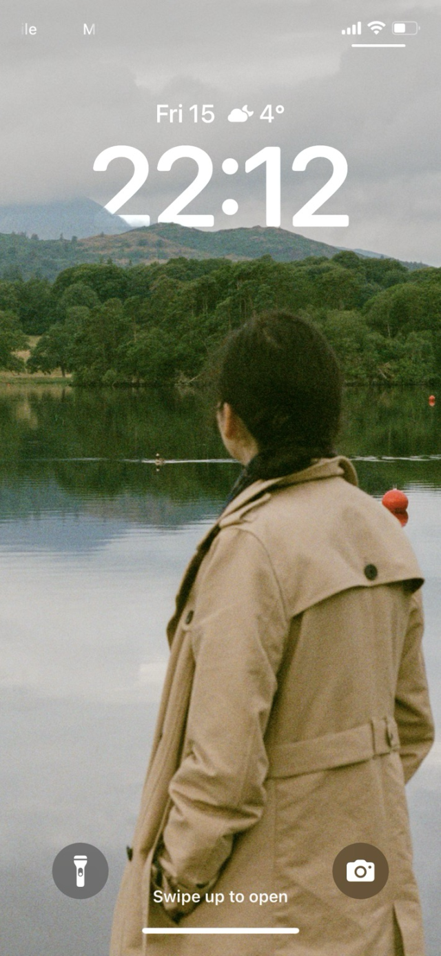 An iPhone lock screen with my photo of my wife looking out onto Windermere, with the clock peaking out from behind the fells in the distance. 