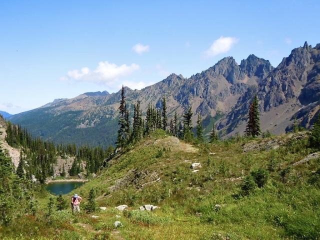  hiker with a red shirt and blue hat walks  uphill from a round blue lake through a green valley.  The background is dominated by ragged dark  brown peaks.