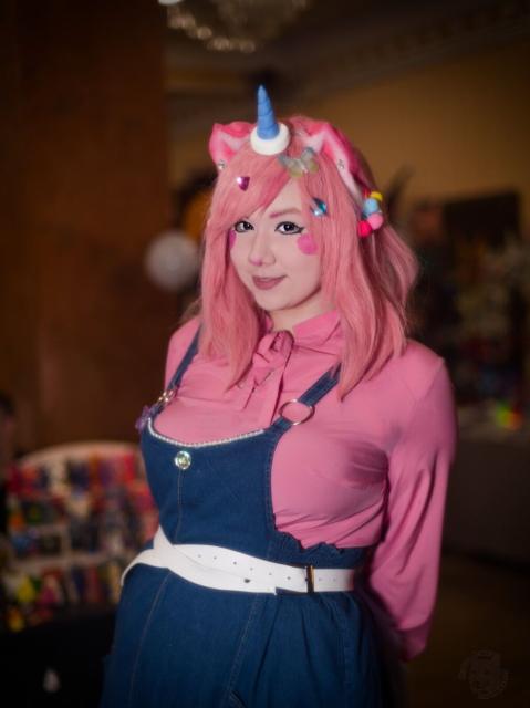 A portrait  of a young woman wearing a pink shirt with long sleeves and a long jeans skirt with a piece of cloth supporte4d by straps that covers chest. Her shoulders-length hair is pink. She wears pink ears and a short blue horn. She holds both her hands behind her back. She looks into the camera and smiles.