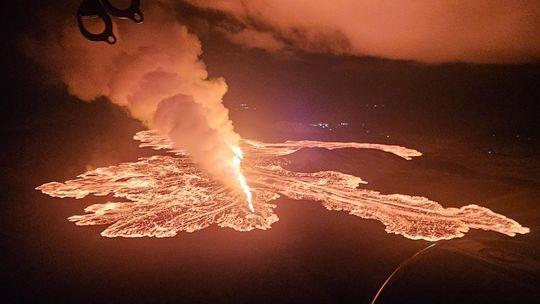 Bright orange lava flowing across dark landscape. A bright line in the centre shows the fissure where lava is erupting. A plume of steam/gases rises from it. Photo by Björn Oddsson / Civil Protection.